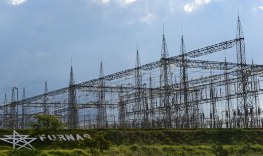 Entrada de estação de geração de energia com destaque para o letreiro à esquerda "Furnas" e torres de transmissão ao fundo