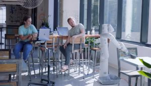 Dois funcionários do Google em refeitório da empresa, sentados em cadeira, vendo um dos robôs fazendo a limpeza