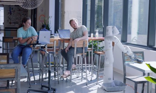 Dois funcionários do Google em refeitório da empresa, sentados em cadeira, vendo um dos robôs fazendo a limpeza