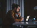 Homem com cabelo no estilo rabo de cavalo pensando em frente ao laptop, alusivo aos investimentos com alta da inflação