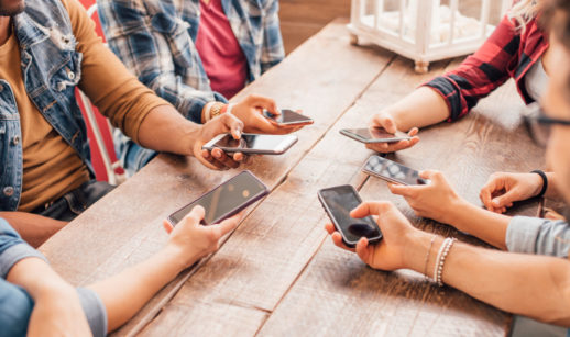 Pessoas com celulares nas mãos juntas sobre mesa de madeira, alusivo à entrada de novos participantes no mercado com o leilão do 5G