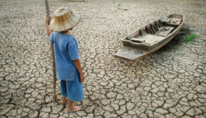 Criança de costas com cajado em frente a barco simples na terra seca, alusivo às mudanças climáticas