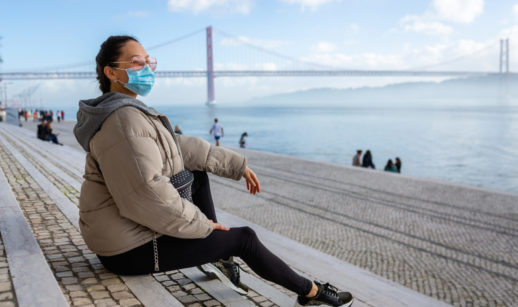 Mulher sentada com casaco bege, de máscara e óculos de sol, olhando a paisagem em Portugal