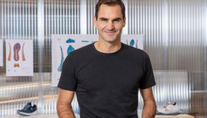 Roger Federer, de camiseta preta, em pé, segurando pares de tênis da On