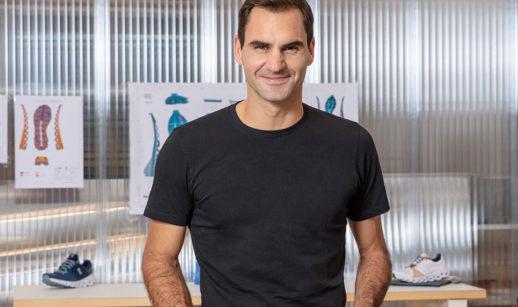 Roger Federer, de camiseta preta, em pé, segurando pares de tênis da On