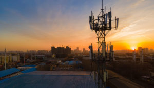 Torre de telefonia móvel em primeiro plano, com paisagem de cidade e pôr do sol ao fundo, alusivo à venda de ativos da Oi
