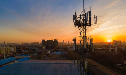 Torre de telefonia móvel em primeiro plano, com paisagem de cidade e pôr do sol ao fundo, alusivo à venda de ativos da Oi
