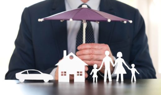Pessoa segurando guarda-chuva de brinquedo pequeno sobre bonecas de famílias, casa e carro, alusivo ao seguro de vida