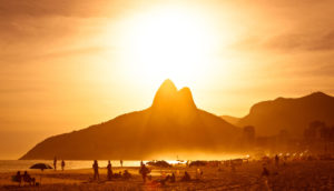 Praia de Ipanema, no Rio de Janeiro, com o Morro Dois Irmãos ao fundo, com sol raiando, alusivo à prevenção ao câncer de pele