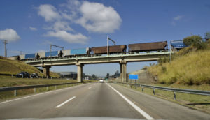 Vista de dentro de carro para trem sobre rodovia, alusivo ao avanço das ferrovias no Brasil
