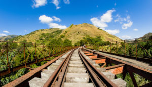 Linha de trem com morro verde e céu azul ao fundo, alusivo ao marco ferroviário