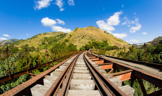 Linha de trem com morro verde e céu azul ao fundo, alusivo ao marco ferroviário