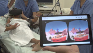 Tablet em primeiro plano mostrando imagens que a criança, em segundo plano, deitada na maca com óculos de realidade aumentada está vendo, alusivo ao metaverso
