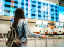 Mulher de costas com mochila e mala de carrinho olhando o painel de voos de aeroporto