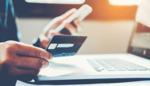 Pessoa segura um cartão de crédito e um celular em frente a um computador, alusivo as compras online