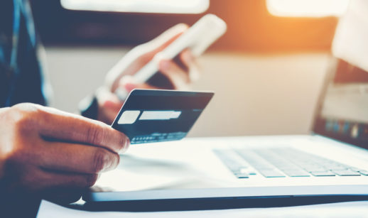 Pessoa segura um cartão de crédito e um celular em frente a um computador, alusivo as compras online