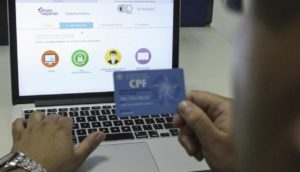 Mãos de pessoa com relógio de pulso prata na esquerda e o cartão de CPF azul na direita inserindo dados no Cadastro Positivo da Serasa Experian