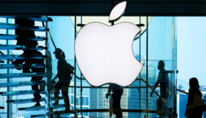 Foto do logo da maçã da Apple, que teve lucro, brilhante na luz branca, com silhueta de pessoas subindo escadas atrás