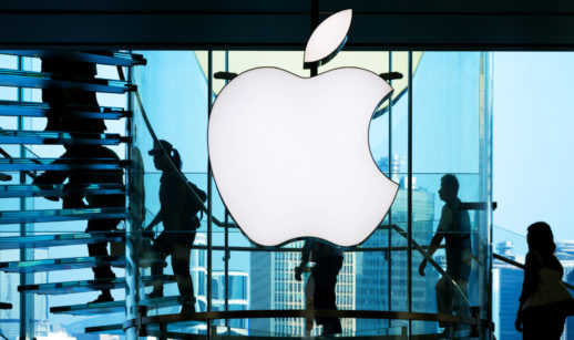 Foto do logo da maçã da Apple, que teve lucro, brilhante na luz branca, com silhueta de pessoas subindo escadas atrás