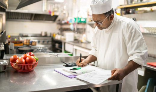 Chef de cozinha parado com caneta, papel e calculadora em mãos, alusivo à contribuição do MEI