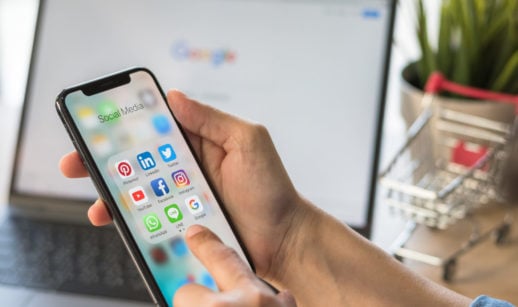 Pessoa segurando iPhone com a tela aberta na pasta de apps de redes sociais e laptop ao fundo desfocado aberto na página do Google, alusivo à proteção de dados pessoais