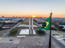 Aérea da Praça dos Três Poderes e da Esplanada dos Ministérios, em Brasília, com bandeira do Brasil em primeiro plano hasteada e pôr do sol ao fundo, alusivo ao Governo Central