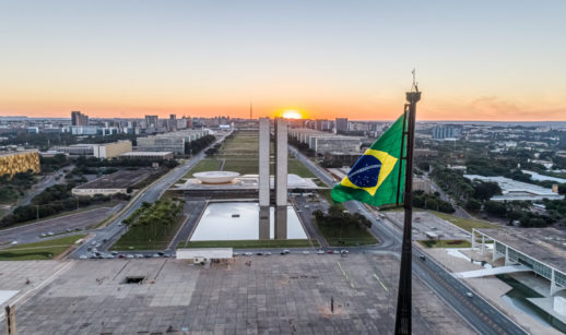 Aérea da Praça dos Três Poderes e da Esplanada dos Ministérios, em Brasília, com bandeira do Brasil em primeiro plano hasteada e pôr do sol ao fundo, alusivo ao Governo Central