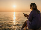 Mulher de costas sentada com celular em mãos e o mar com pôr do sol ao fundo, alusivo aos investimentos para janeiro