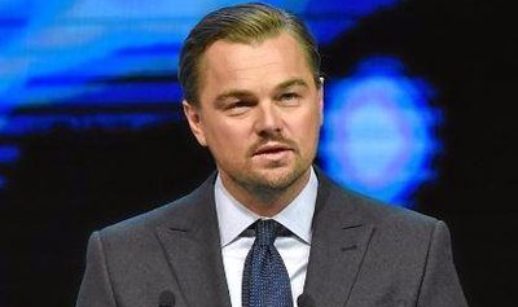 Leonardo DiCaprio, de terno cinza e gravata azul, em frente a microfones, falando