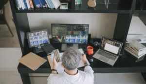 Vista de cima de investidor com cabelos grisalhos curtos analisando telas de computador e com papel nas mãos, alusivo ao mercado de opções