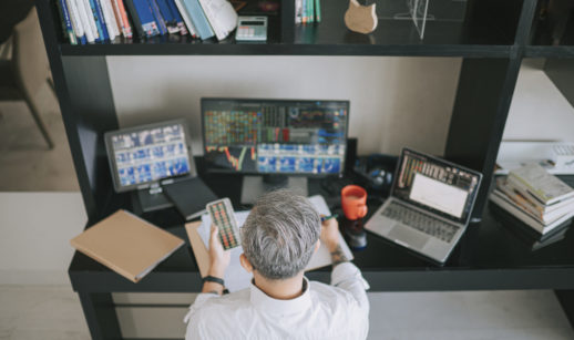 Vista de cima de investidor com cabelos grisalhos curtos analisando telas de computador e com papel nas mãos, alusivo ao mercado de opções