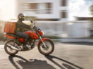 Uma das motos de delivery, na cor vermelha, rodando em rua com motociclista e bolsa de comida nas costas