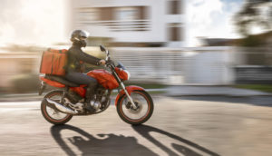 Uma das motos de delivery, na cor vermelha, rodando em rua com motociclista e bolsa de comida nas costas