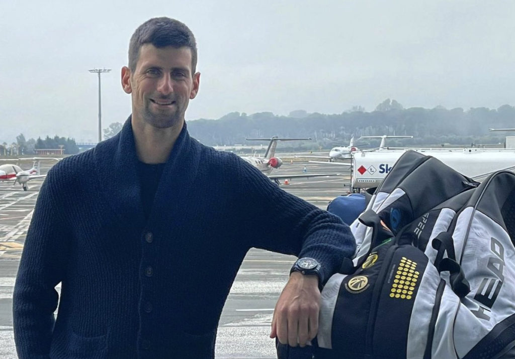 Novak Djokovic, tenista sérvio com suéter preto e braço esquerdo apoiado sobre monte de malas em aeroporto