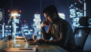 Homem de suéter bege sentado em frente a laptop pensando e ao fundo a cidade iluminada, alusivo às operações a termo do mercado financeiro