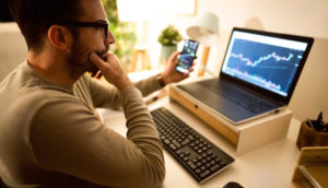 Homem com óculos e suéter bege olhando para laptop e celular com gráficos de operações de renda variável