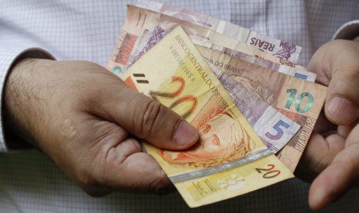 Mãos de pessoa contando notas de 5, 10 e 20 reais, alusivo ao serviço de consulta junto aos bancos