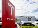 Totem vermelho com letreiro e logo da Tesla, que teve lucro, em branco, em frente a uma concessionária com veículos estacionados