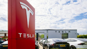 Totem vermelho com letreiro e logo da Tesla, que teve lucro, em branco, em frente a uma concessionária com veículos estacionados