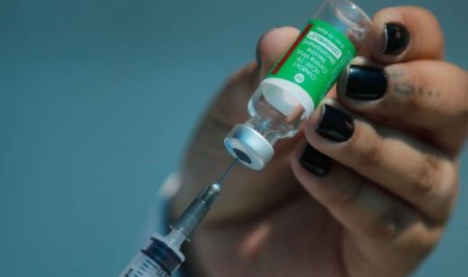 Close de mãos com unhas pintadas de preto puxando com seringa a dose da vacina da Fiocruz contra a covid-19