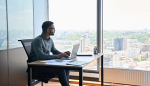 Homem de camisa social azul, sentado, com laptop na mesa, olhando pela janela de um prédio, alusivo aos investimentos em fevereiro