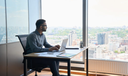 Homem de camisa social azul, sentado, com laptop na mesa, olhando pela janela de um prédio, alusivo aos investimentos em fevereiro