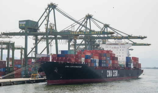 Navio nas cores preta e vermelha com contêineres ao lado de berço com pontes rolantes no terminal de Santos, um dos portos do setor portuário do Brasil