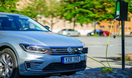 Carro da Volkswagen na cor prata abastecendo com energia elétrica, com dia ensolarado e árvores verdes ao fundo desfocadas
