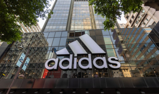 Fachada de loja da Adidas em prédio espelhado, com destaque para o logo em branco e árvores próximas nas laterais