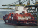 Navio cargueiro atracado no Porto de Santos e sendo carregado por pontes rolantes verdes, alusivo à balança comercial brasileira de fevereiro