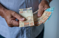 Close em mãos de uma pessoa com camisa azul contando notas de 50 e 100 reais, alusivo à restituição do IRPF 2022
