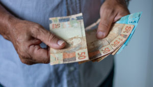 Close em mãos de uma pessoa com camisa azul contando notas de 50 e 100 reais, alusivo à restituição do IRPF 2022