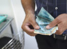 Close em mãos de homem com camisa azul e calça preta conntando notas de 100 reais, alusivo à Ume, que fornece crédito