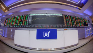 Interior da B3, com destaque para balcão com microfone e logo da empresa e telão de LED gigante com informações sobre o mercado financeiro, alusivo às top picks para investir em renda variável na semana